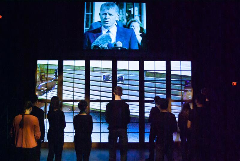 A group of people stands in front of a large screen composed of multiple smaller panels. The screen displays an old television image of a man speaking at a podium. The scene is indoors with dim lighting.