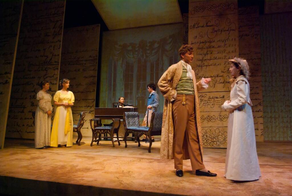 A theater scene with actors in period costumes. Two women in long dresses and bonnets stand on the right. A man in a coat talks to a woman. Another man sits at a desk in the background, with script-covered walls surrounding the stage.