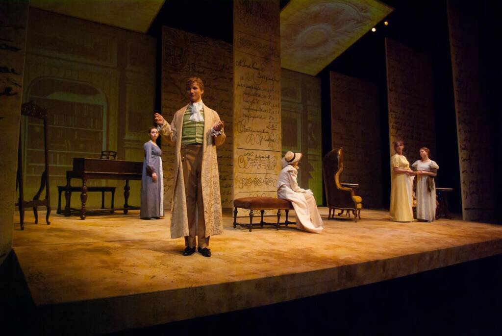 A theatrical stage with actors dressed in historical costumes. A man in a beige coat speaks to the audience, while others in period attire stand and sit in the background. The set features large panels with writing and a desk with chairs.