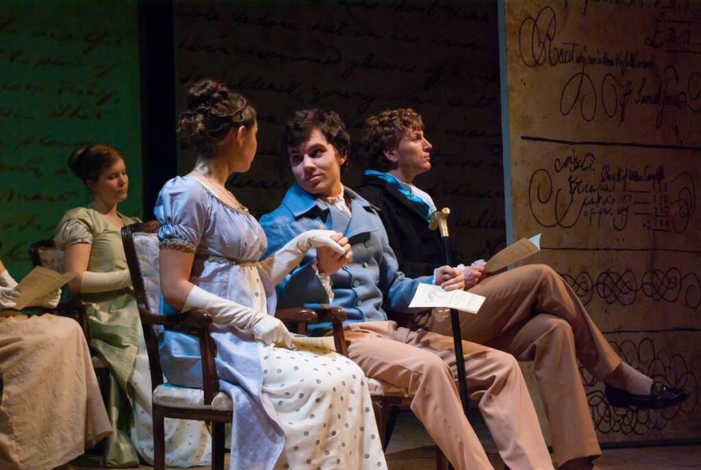 Four people in period costumes sit on stage. A woman in a white dress faces a man in a blue jacket, holding hands. Two others sit nearby, one with a cane. The backdrop features cursive writing.