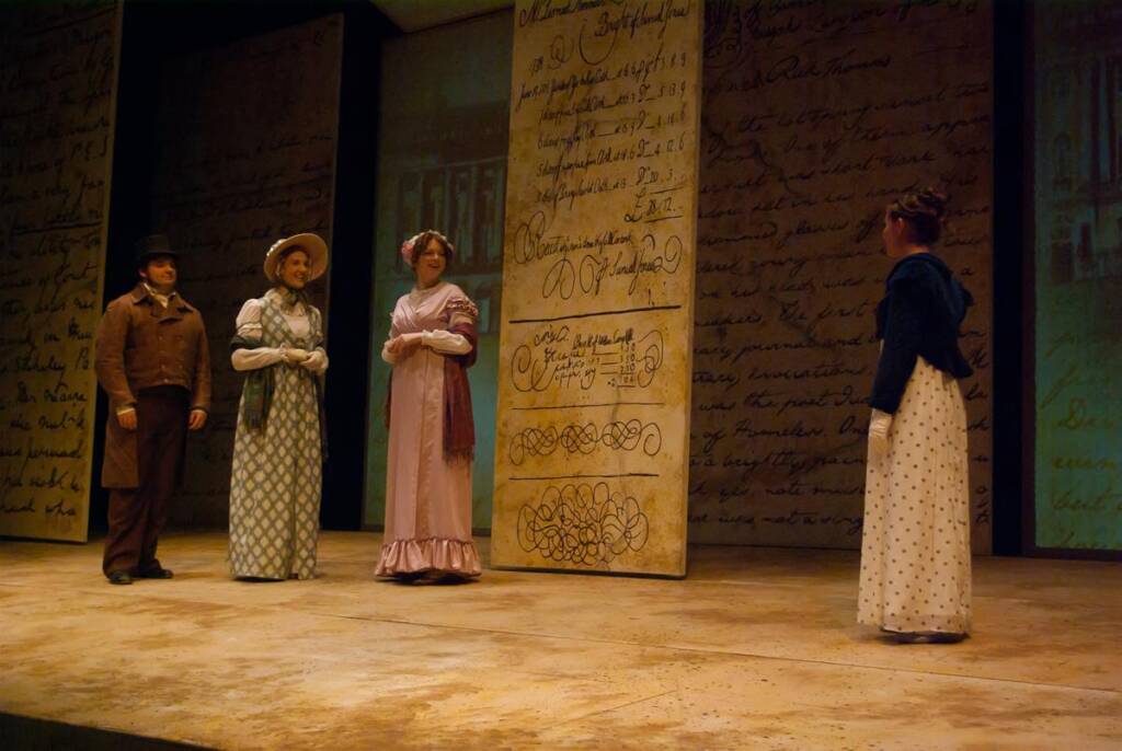 Four people in period costumes stand on stage, with a backdrop of large handwritten letters. Two women in long dresses face another woman in a similar dress. A man wearing a brown coat and hat stands beside them. The scene has a historical setting.