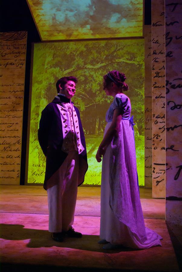 A man and woman in 19th-century attire stand facing each other on a stage. The background features images of handwritten letters and a scenic landscape. The lighting casts a warm glow, creating a historic, romantic atmosphere.