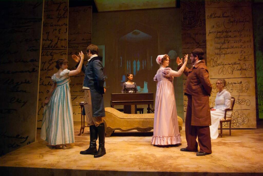 A group of actors in period costumes perform a scene on stage. Two pairs dance while others watch. The stage is decorated with large, scripted papers and a backdrop with dim lighting, creating a historical atmosphere.
