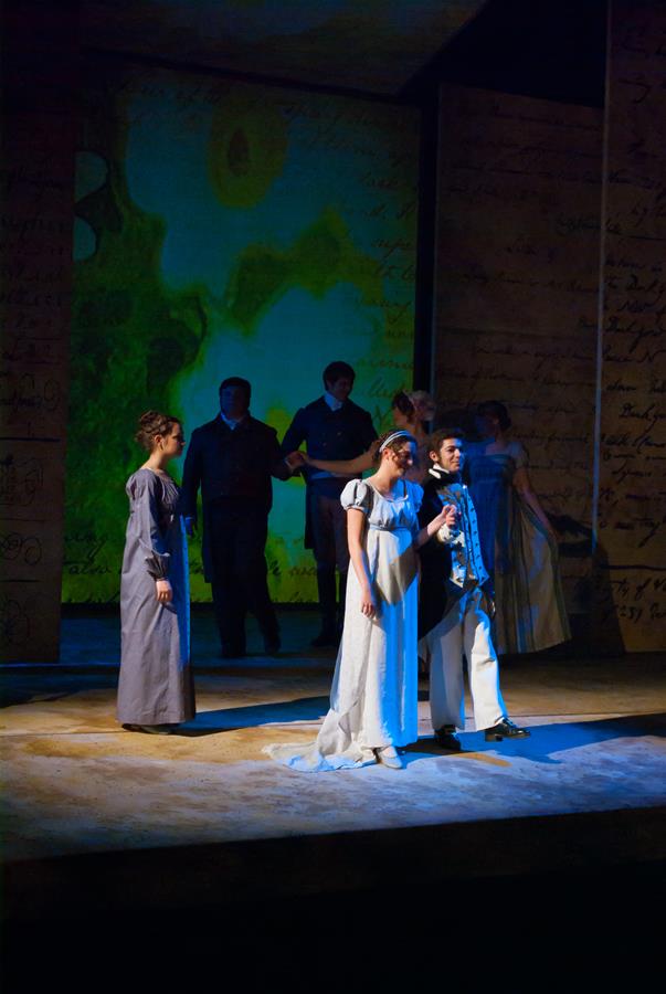 A theatrical performance on stage with actors in period costumes. One actor in a white dress and another in a dark suit stand together in the spotlight, with other actors in the background. The backdrop features abstract art and script.