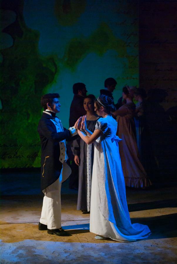 A group of people dressed in period costumes are dancing in a dimly lit room. The focus is on a man and woman facing each other with their hands raised, as if about to waltz. Others are seen dancing in the background. The ambience is historical.