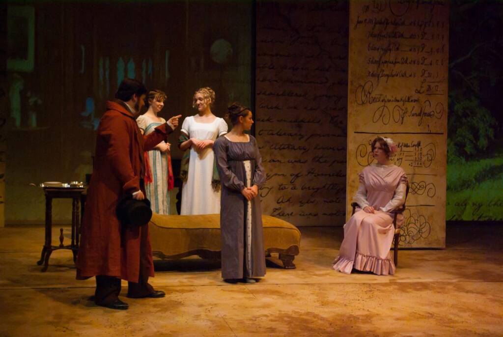 A theatrical scene with five actors in period costumes. Three women stand in long dresses, one points toward a seated woman in a pink gown. A man in a red coat and hat stands nearby. The backdrop features large handwritten text.