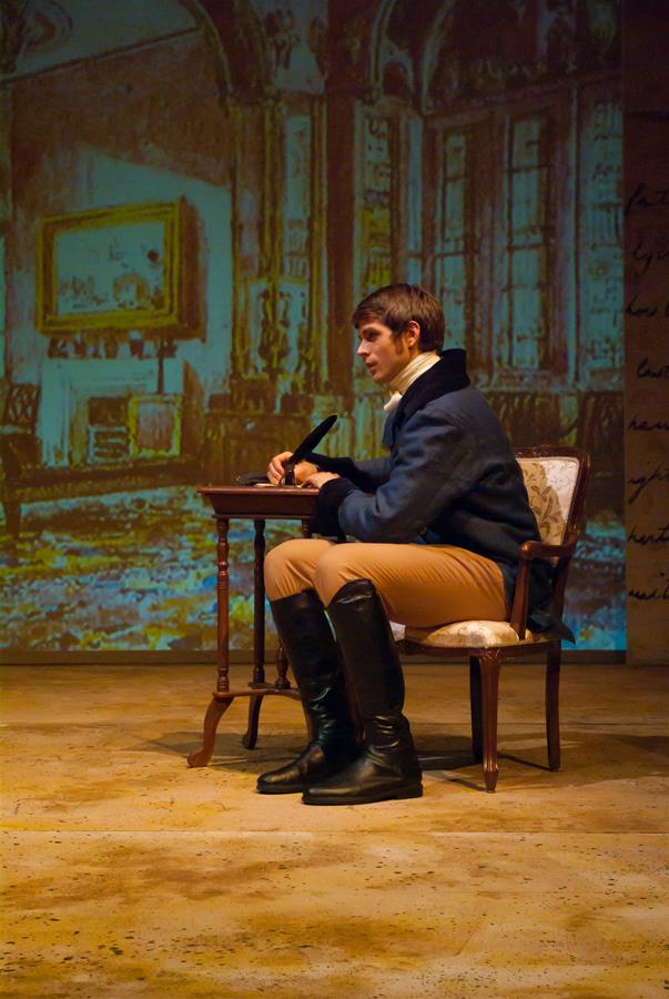 A person in period costume sits at a small table, writing with a pen. They wear a dark jacket, beige pants, and black boots. The background shows a projected image of a vintage room with large windows and ornate decor.