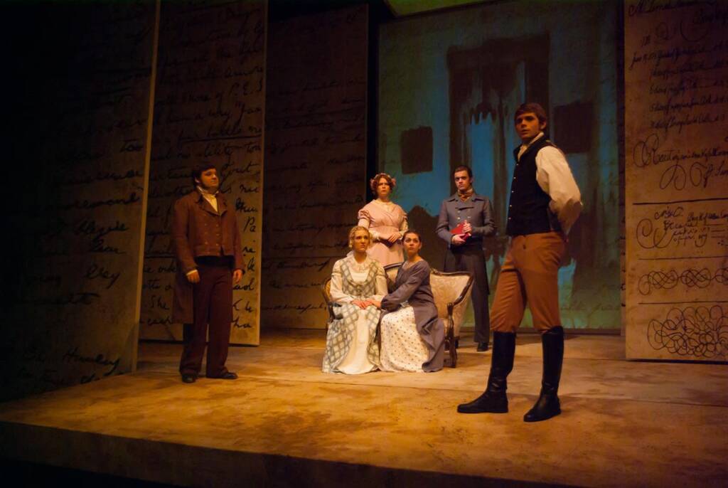 A theater scene with six actors dressed in period costumes. Two women sit in the center, surrounded by four standing men. The backdrop features large handwritten letters in dim lighting.