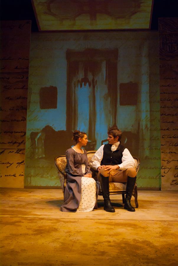 A woman in a long dress sits on a vintage sofa next to a man in historical costume attire. They are in a theatrical setting with a projected backdrop featuring a large shadowy door and script-like text.