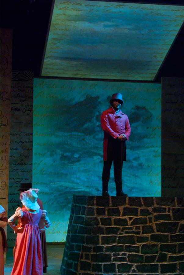 A theatrical scene with a man in a red coat and hat standing on a stone platform, illuminated against a backdrop that appears to have script and an abstract image. Two people in period costumes are on the left.