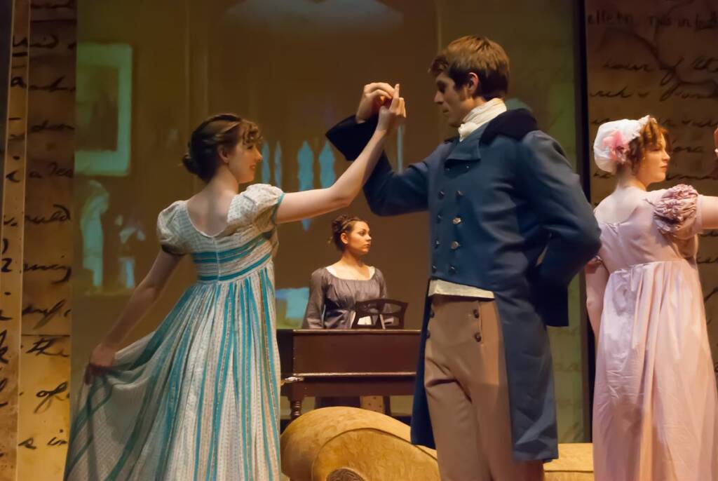 Actors in Regency-era costumes perform on stage. A man in a blue coat and a woman in a flowing dress dance gracefully. Another woman in pink stands nearby, while a person in the background plays the piano. The backdrop features vintage writing.