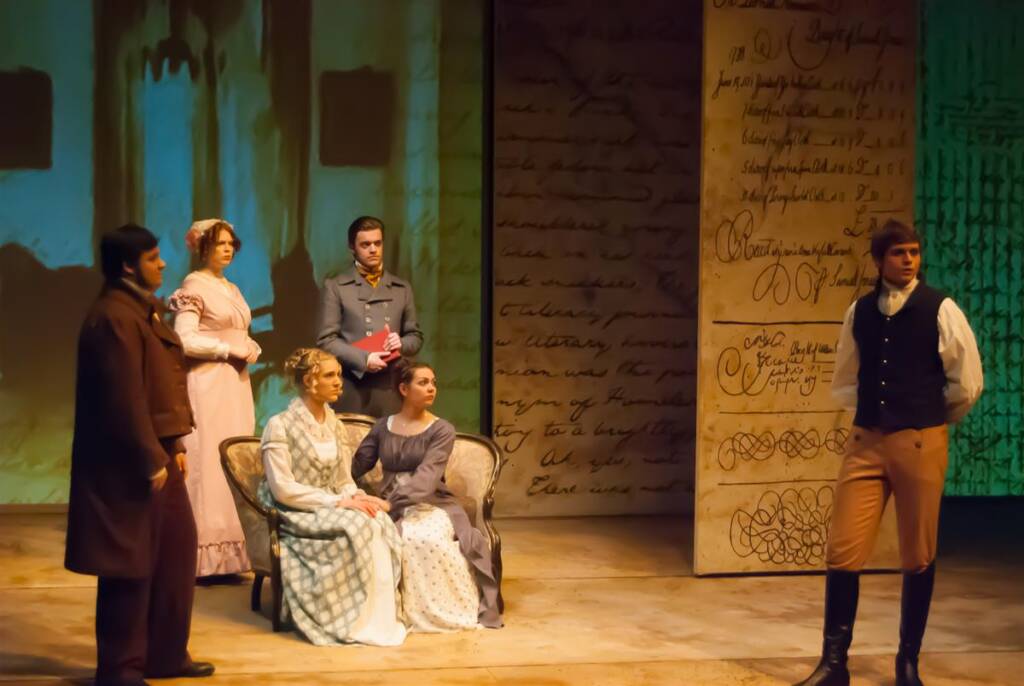 A group of six actors in period costumes perform on stage. Two men and two women are seated, while a man and woman stand. The background displays large handwritten text. The setting suggests a historical drama.