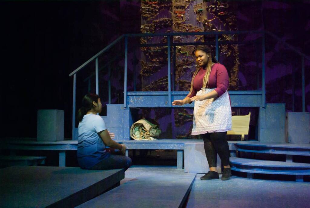 Two women are on a dimly lit stage with an abstract backdrop. One, standing, wears a pink top and apron, gesturing expressively. The other, sitting on a step, listens intently wearing a white top. A large bag lies nearby.