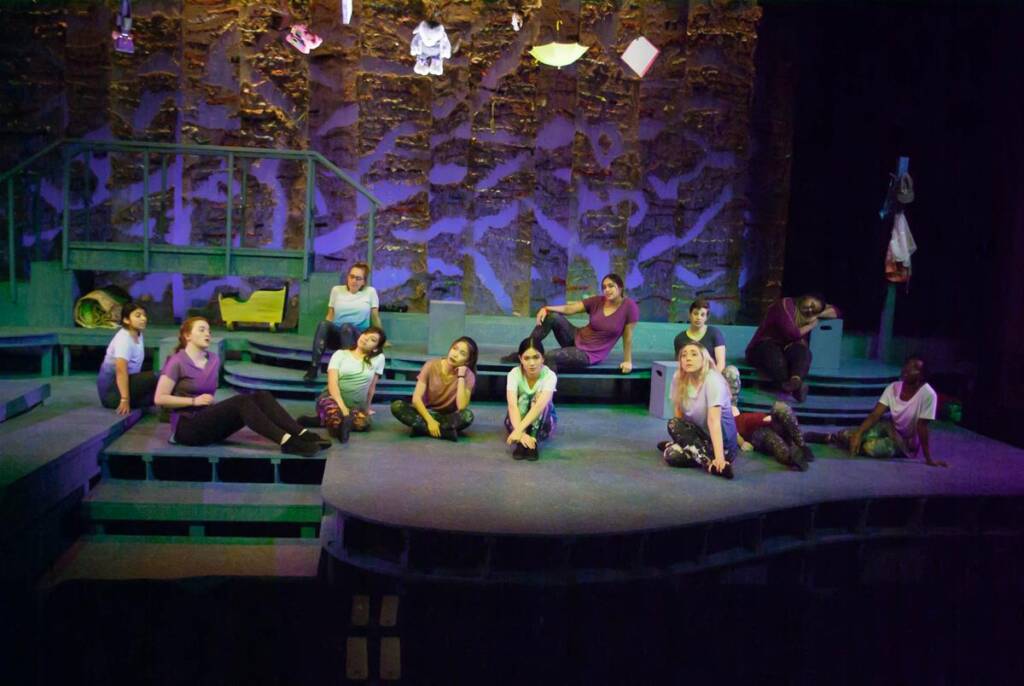 A group of performers sits and lounges on a multi-level stage with a textured stone-like backdrop. They are dressed in casual, colorful attire. Hanging objects, including a small umbrella, are visible above them. The lighting casts a moody, dramatic effect.