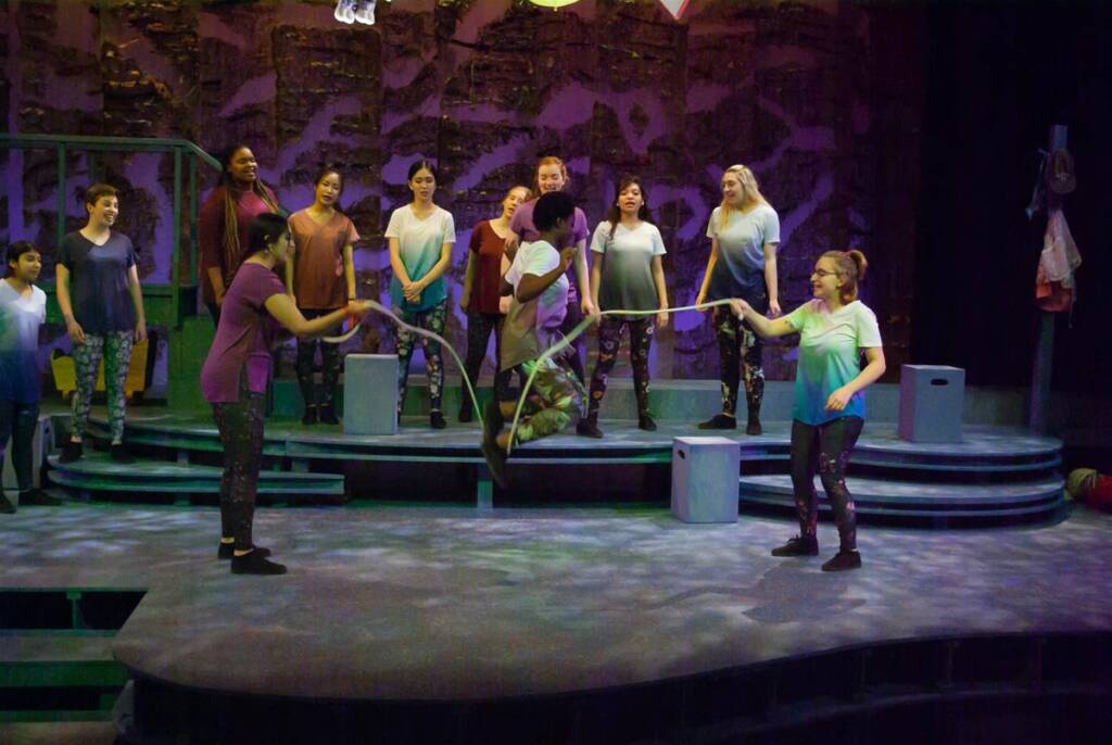 A group of people on stage, some holding a jump rope while one person jumps. Others stand and watch. They're in casual attire with colorful lighting and a backdrop resembling a rocky surface. The scene gives a lively and engaging atmosphere.