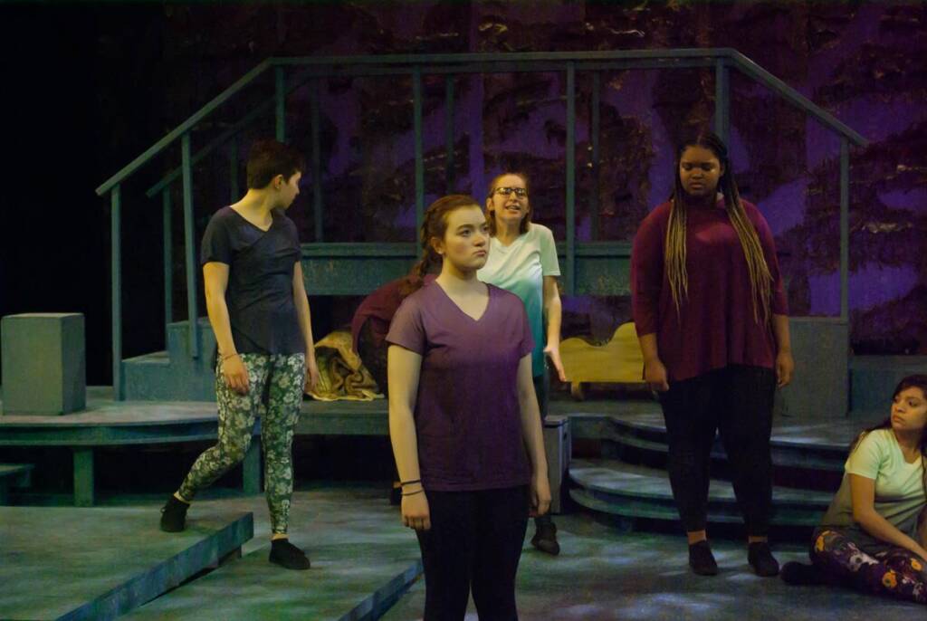 A group of five people is on a dimly lit stage. The central figure is a person with a focused expression, wearing a purple shirt. Others stand or sit around, engaged. The setting has a muted, theatrical backdrop with steps and abstract decor.