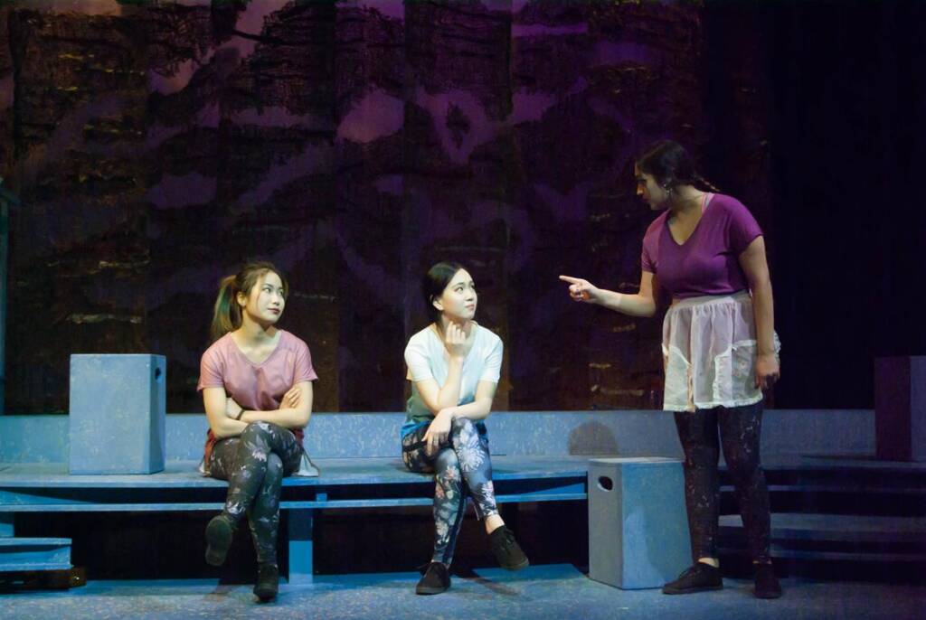 Three actors on stage: two seated on blue platforms, one standing. The standing actor, in a purple top and apron, gestures with a finger, while the seated actors look attentive. The background is dark with textured, abstract scenery.