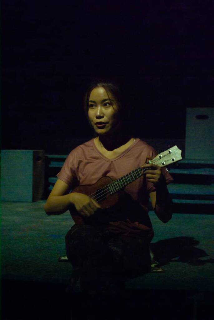 Person sitting in dim light, holding a ukulele. They are wearing a pink shirt and looking slightly to the side. The background is dark and indistinct.