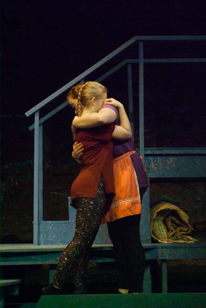 Two people are embracing on a stage with a dark background. They stand near blue stairs. One wears a reddish-brown top and patterned leggings, and the other is in a purple top and orange skirt.