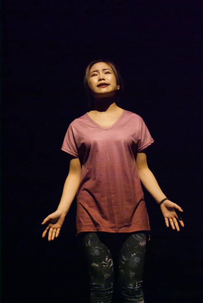 A woman stands on a dark stage, wearing a maroon t-shirt and patterned leggings. Her arms are slightly extended, and she appears to be singing or acting, with an expressive face illuminated by stage lighting.