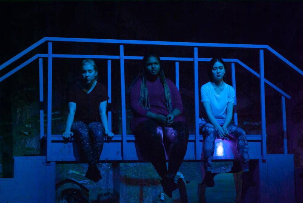 Three people sit on a bridge-like structure on stage, illuminated by blue lighting. They are spaced evenly and appear to be in a performance or rehearsal setting. The atmosphere is dramatic and focused.