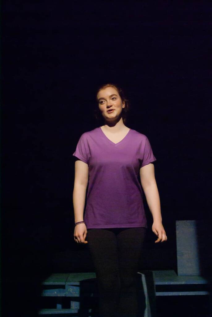 A person in a purple shirt and dark pants stands on stage in a spotlight against a dark background. They appear to be speaking or performing.