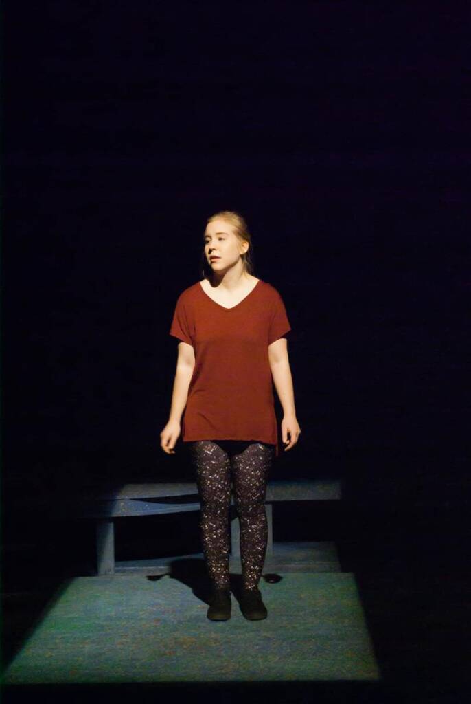 A person stands alone on a dimly lit stage, wearing a maroon shirt and patterned leggings. They are illuminated by a spotlight, with their hands by their sides, while a bench is visible behind them.