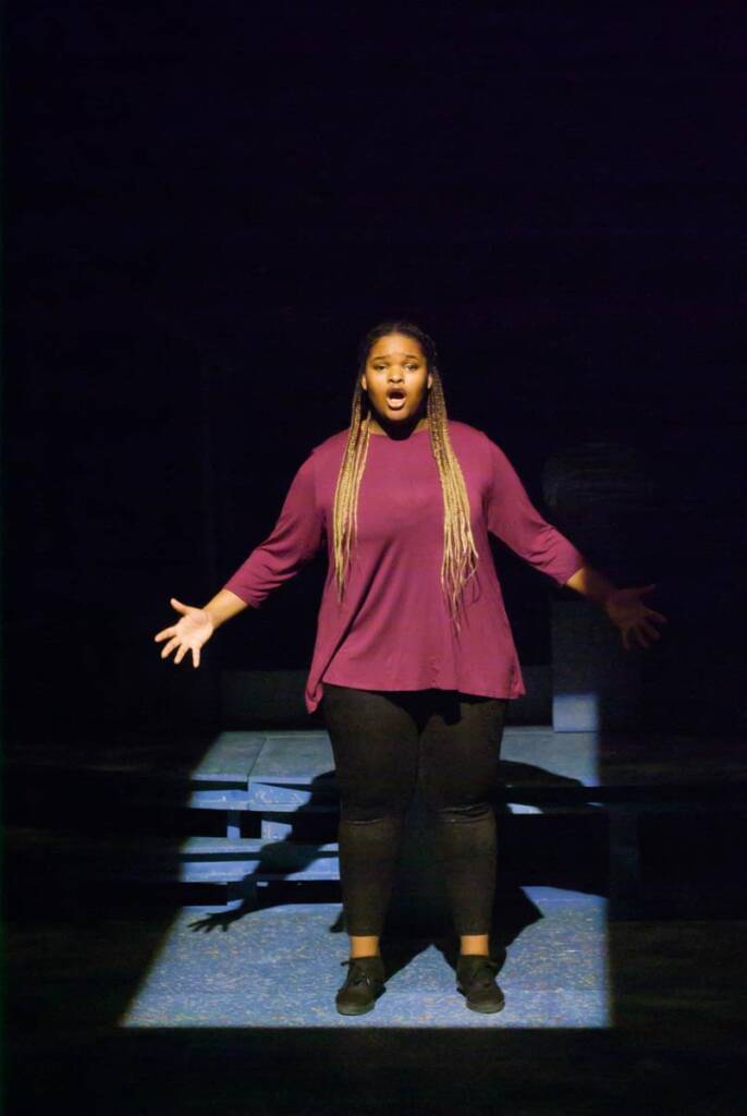 A woman with long braided hair stands on a dimly lit stage, wearing a purple top and black pants. She appears to be singing or performing passionately, with her arms slightly extended.