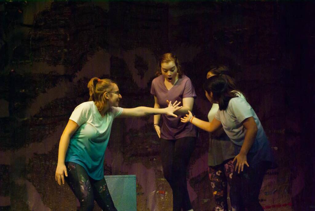 Four performers on stage act out a scene. One woman reaches out dramatically while others react. They are in casual clothing, and the background is dimly lit, suggesting an intense or emotional moment in a play.