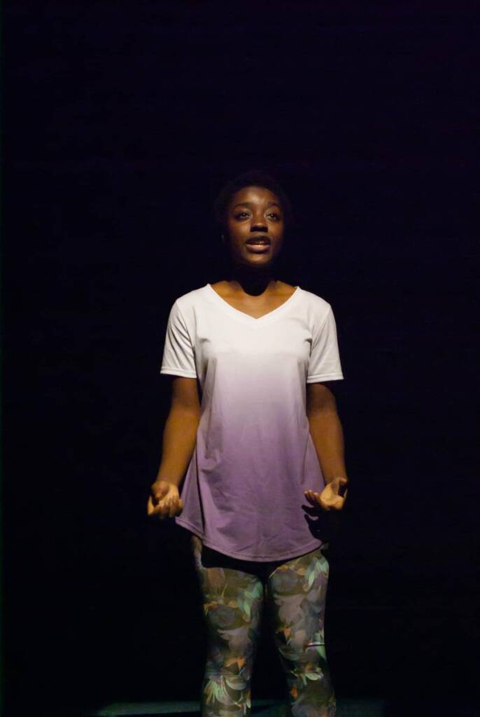 A person stands on stage against a dark background, wearing a white gradient t-shirt and patterned pants. Their hands are slightly raised, and they appear to be speaking or singing.