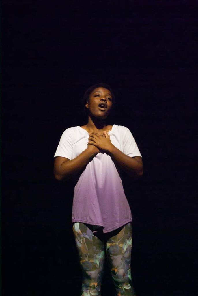 A person stands on a dark stage with eyes closed and hands clasped over their chest. They wear a white shirt and patterned pants, illuminated by a spotlight.