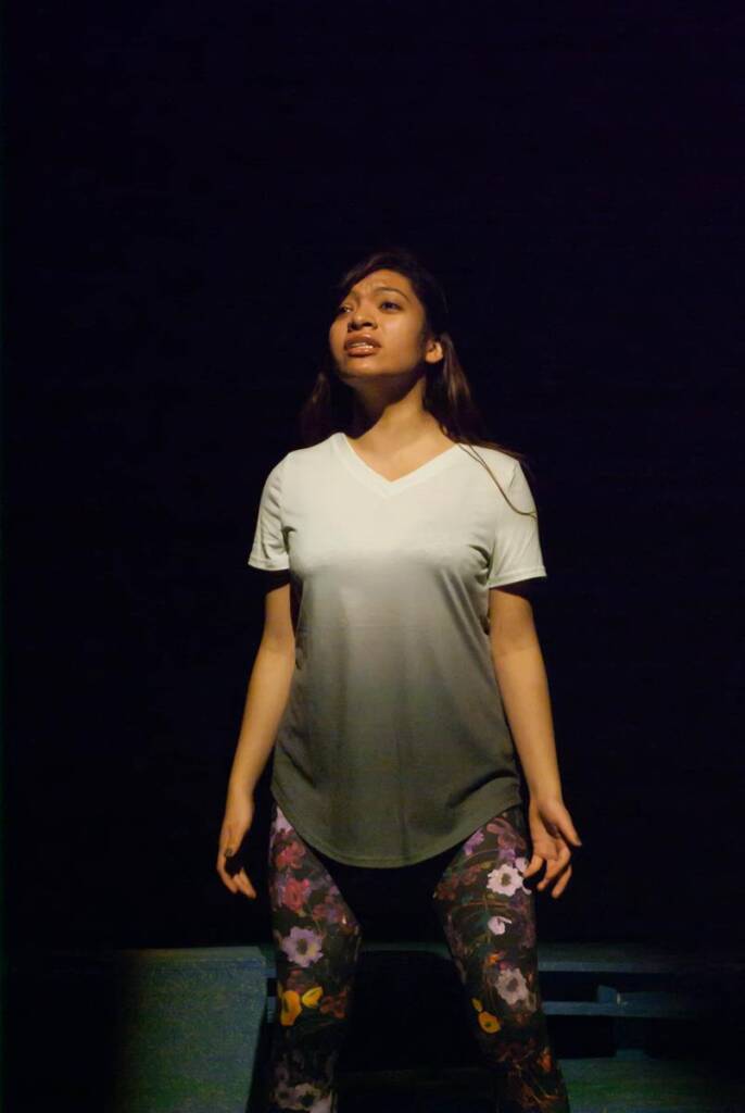 A person stands confidently under stage lighting, wearing a light-colored shirt and floral leggings. The background is dark, highlighting their expressive pose.