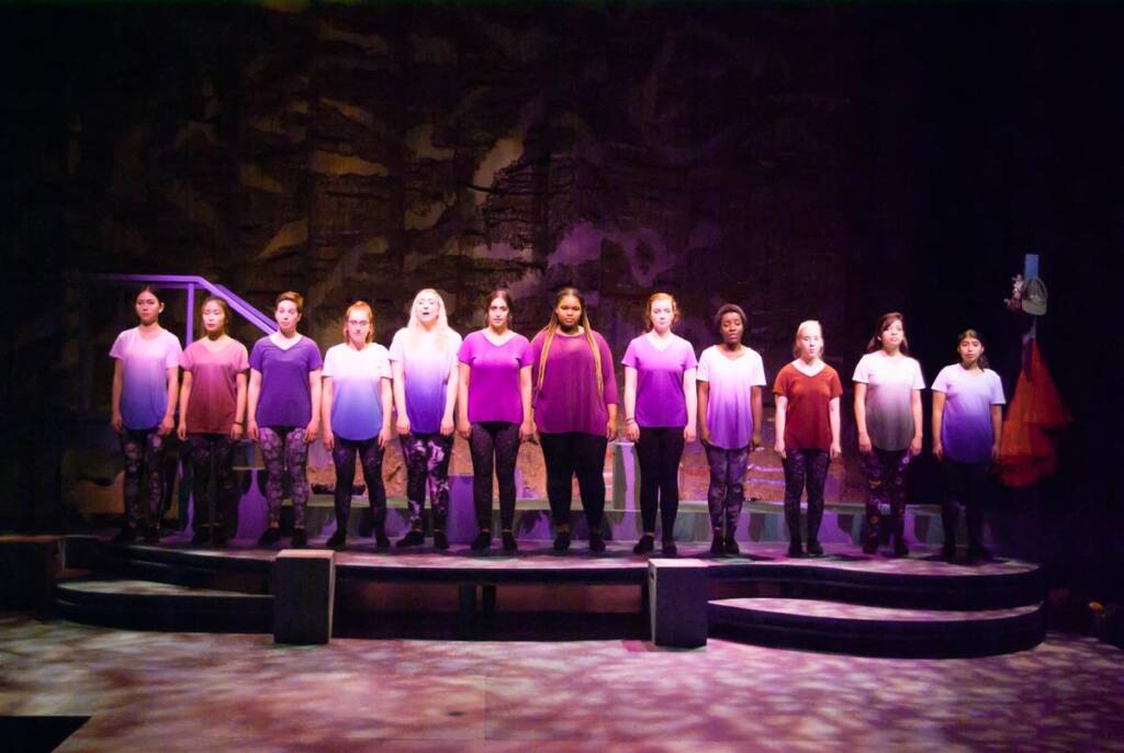 A group of performers stands on stage in coordinated purple and white outfits, varying from light to dark shades. They're lined up in a row, looking forward, with a dark, textured backdrop behind them. The stage floor is patterned with light effects.