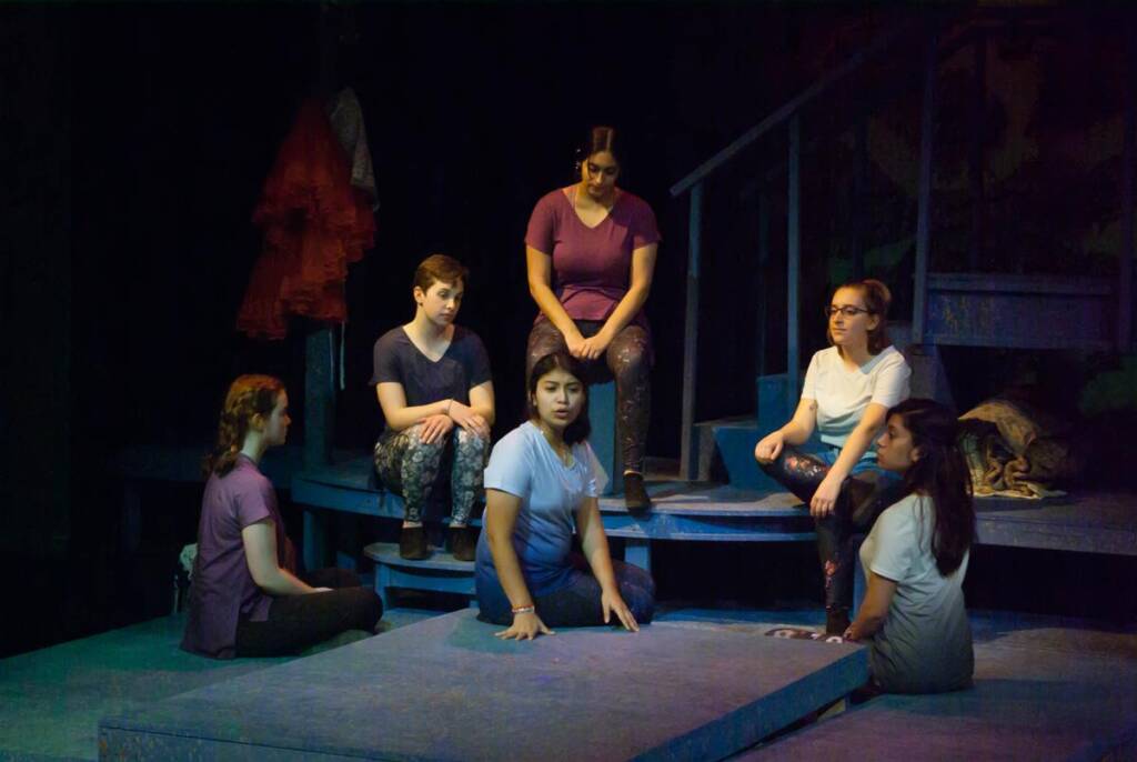 A group of young women sit on a stage in a dark theater, engaged in a conversation. They are casually dressed and seated on blue steps. The spotlight is on them, creating a focused ambiance. A staircase and hanging clothing are visible in the background.