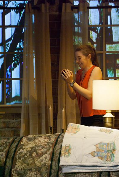 A person in a red sleeveless top holds a small object and smiles by a floral-patterned couch. Behind them are large windows with curtains, through which trees are visible. A lamp and folded quilts are on the couch.
