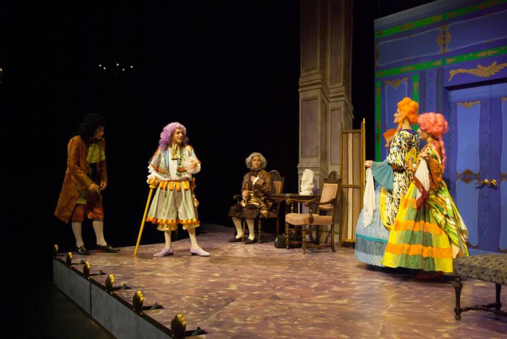 A theatrical scene with four actors in colorful, ornate period costumes on stage. One actor with a purple wig holds a cane. The backdrop includes a blue wall with green and gold trim, and stage props like a chair and table.