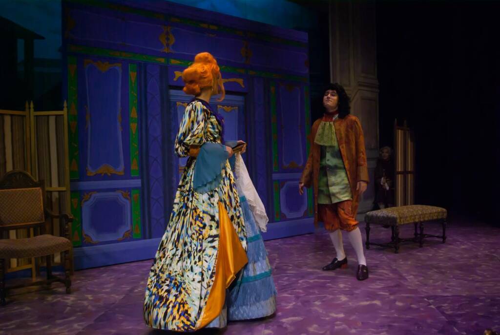 Two actors in a colorful stage play. One wears an elaborate orange wig and a colorful, patterned gown. The other is in a brown coat, green waistcoat, and white stockings. The set features a large, ornate backdrop with purple and gold designs.