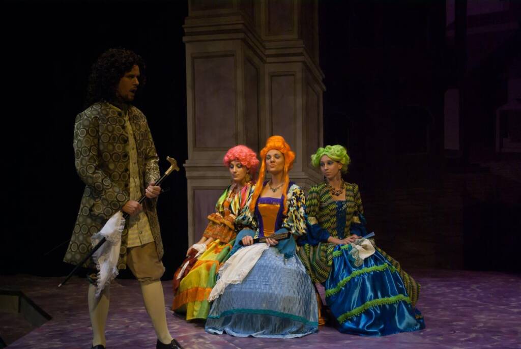 A man in vintage clothing with a cane stands to the left, while three women in colorful 18th-century costumes with elaborate wigs sit on a stage to the right. The backdrop is reminiscent of classical architecture.