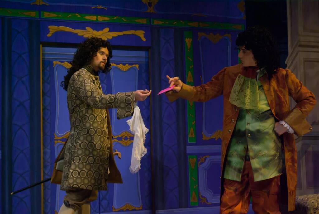 Two actors in historical costumes perform on stage. One in a patterned coat offers a white handkerchief, while the other in an ornate suit and frilled cravat gestures dramatically. The backdrop features a detailed, colorful design.