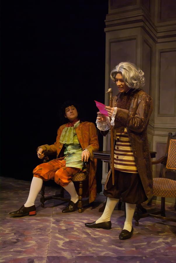 Two actors in period costumes perform on stage. One sits in a chair wearing orange clothing and a wig, while the other stands reading a pink paper, dressed in brown attire and a grey wig. The background is a simple, dark theatrical set.