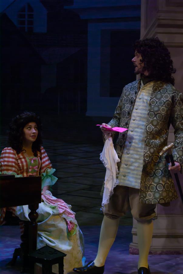 Two actors in period costumes are performing on stage. One is seated, wearing a striped and floral dress, looking upward. The other stands in an ornate jacket, holding a folded paper and a cloth, with a theatrical backdrop behind them.