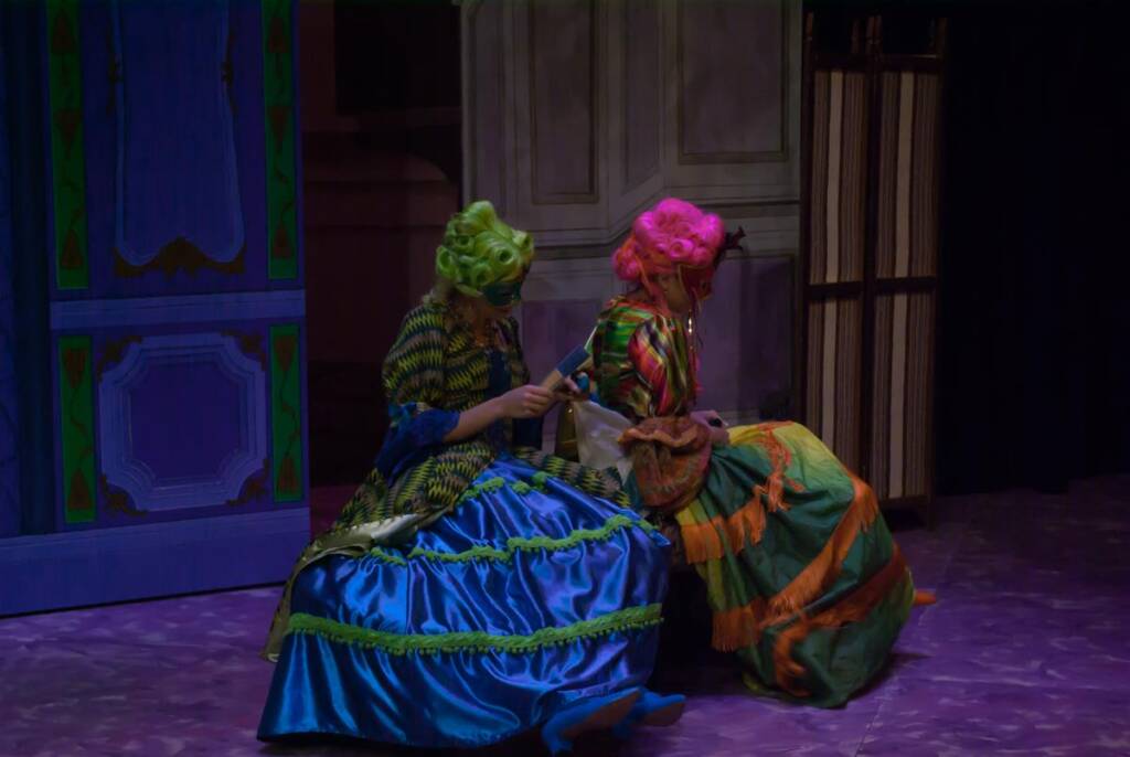 Two people dressed in vibrant, colorful costumes and wigs sit on a stage. One person is in a blue and green dress with a green wig, the other in an orange and green dress with a pink wig, both facing away, with stage props in the background.