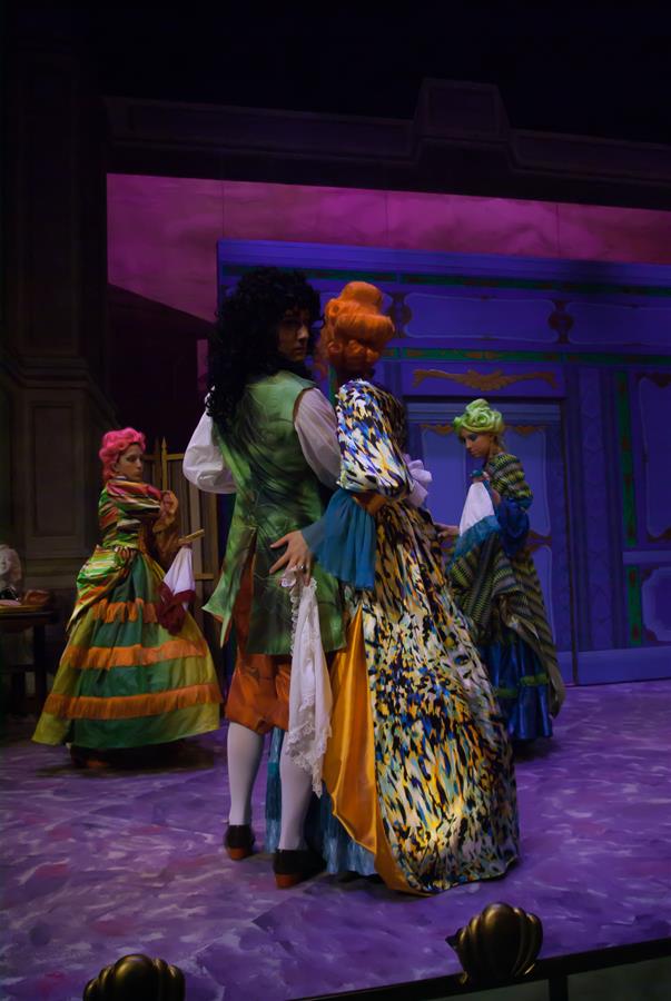 A theatrical scene with actors in colorful, elaborate period costumes. One actor in green and another in a multicolored dress are centered stage, while others in vibrant attire are in the background. The set has a painted, ornate backdrop.