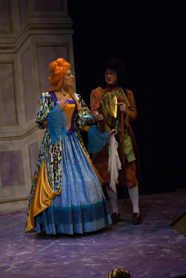 Two actors in period costumes perform on stage. The woman wears a bright dress with orange and blue details, her hair styled elaborately. The man holds a fan and wears a dark hat and coat. They stand against a backdrop resembling ornate walls.