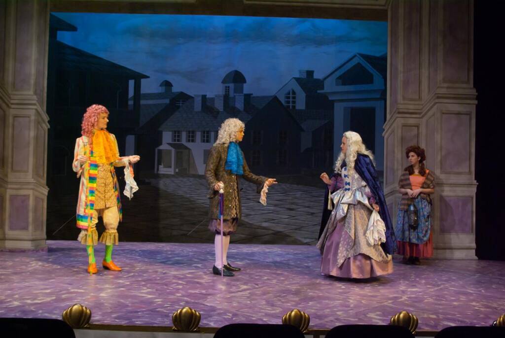 Four actors in elaborate, period costumes perform on a stage, depicting a historical scene with a backdrop of a vintage town. Two actors in wigs dominate the center, gesturing dramatically, while two others watch from the sides.