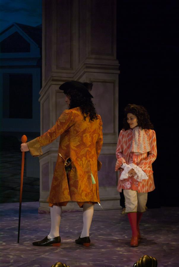 Two actors in period costumes on stage. The person on the left wears a long orange coat, breeches, and a black hat, holding a cane. The person on the right wears a pink patterned coat, white lace, and red boots. The backdrop resembles a formal setting.