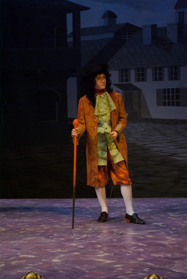 A person stands on a stage dressed in historical attire, including a long wig, brown coat, green waistcoat, breeches, and white stockings. They hold an orange cane. The backdrop features a dark, stylized street scene with buildings.