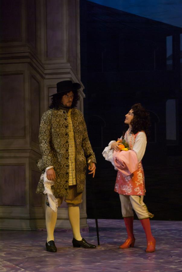 Two actors on stage in historical costumes. The man wears a patterned coat, yellow stockings, and a wide-brimmed hat, while the woman holds flowers and wears a pink outfit with red boots. They stand against a backdrop of a large building.