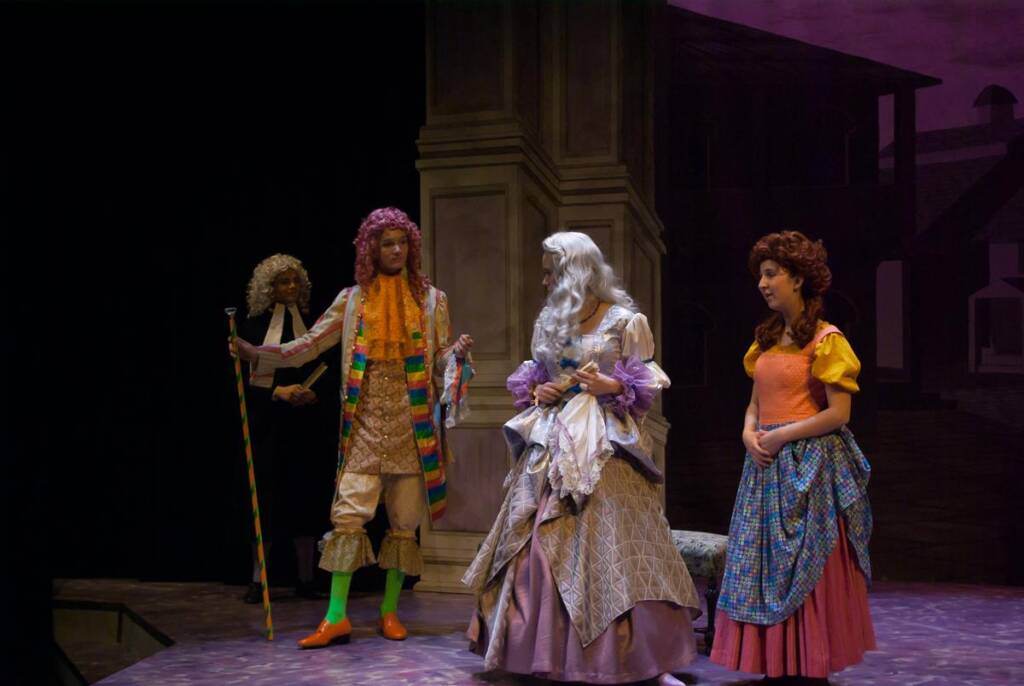 A theatrical scene with four actors in period costumes. One actor wears a long coat and holds a staff, another wears a vibrant outfit with a scarf. A third actor is in a purple gown, and the fourth wears a yellow top with a long skirt, against a stage backdrop.