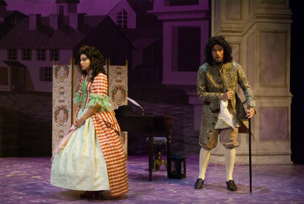 Two actors on stage in period costumes: a woman in a red and white checkered dress with lace trim, and a man in a patterned coat holding a cane. The backdrop features a painted exterior of houses. They appear engaged in a dramatic scene.